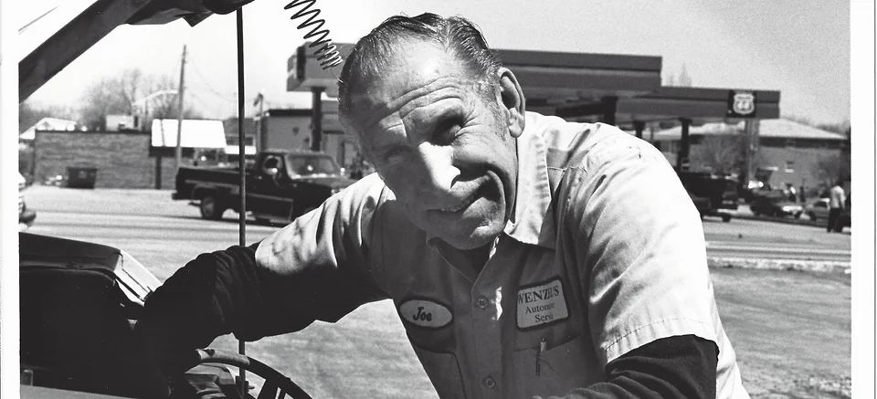 Black and white photo of a Wenzel's Automotive mechanic leaning on a car at the shop