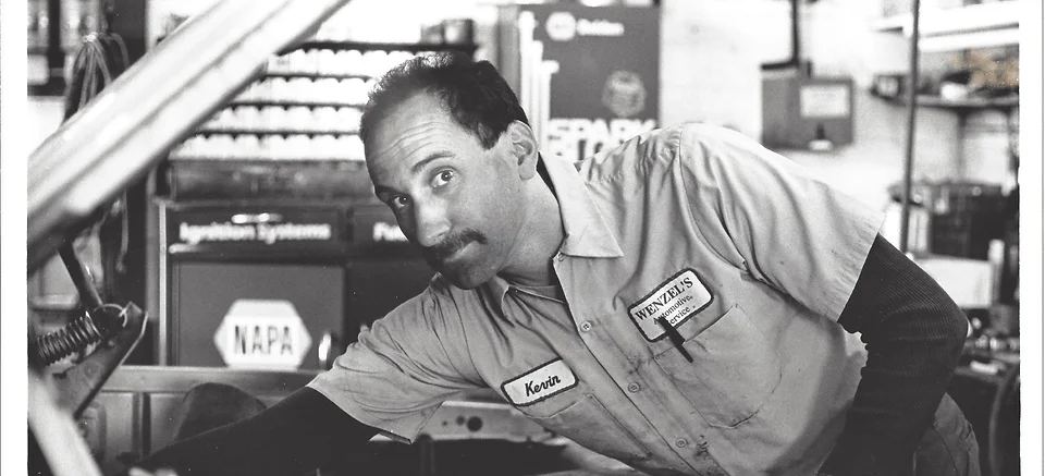Black and white photo of a Wenzel's Automotive technician in uniform leaning over a car in the service bay